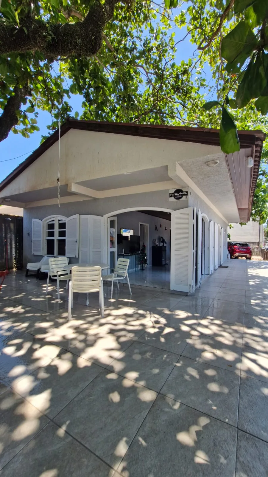 Varanda de canto da casa, com mesa e cadeiras no pátio de porcelanato, sob a sombra da figueira